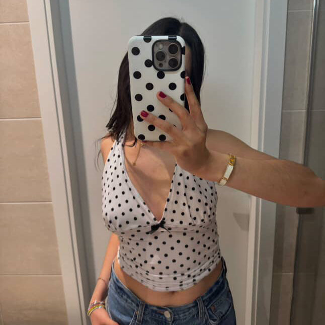 Person taking a mirror selfie wearing a polka dot top and jeans in a bathroom.