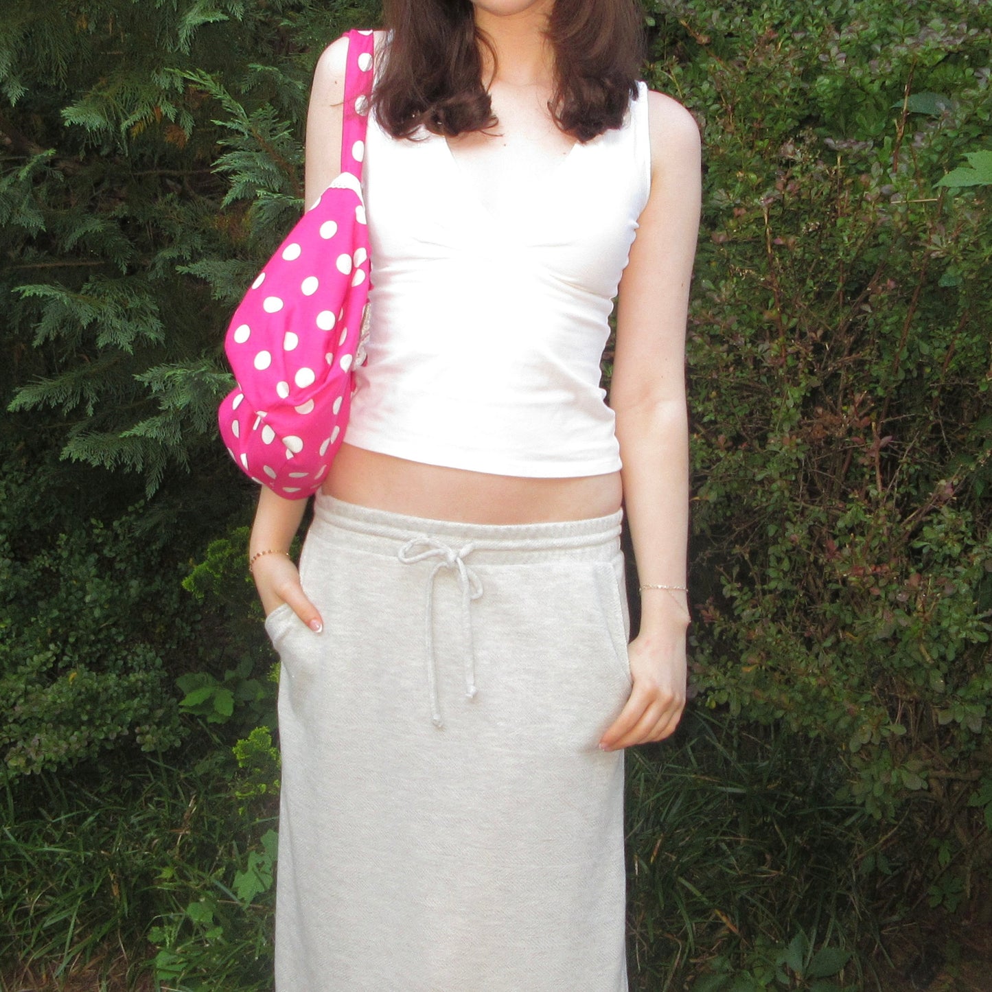 Person wearing a white sleeveless top and gray sweatpants, holding a pink polka dot bag outdoors.