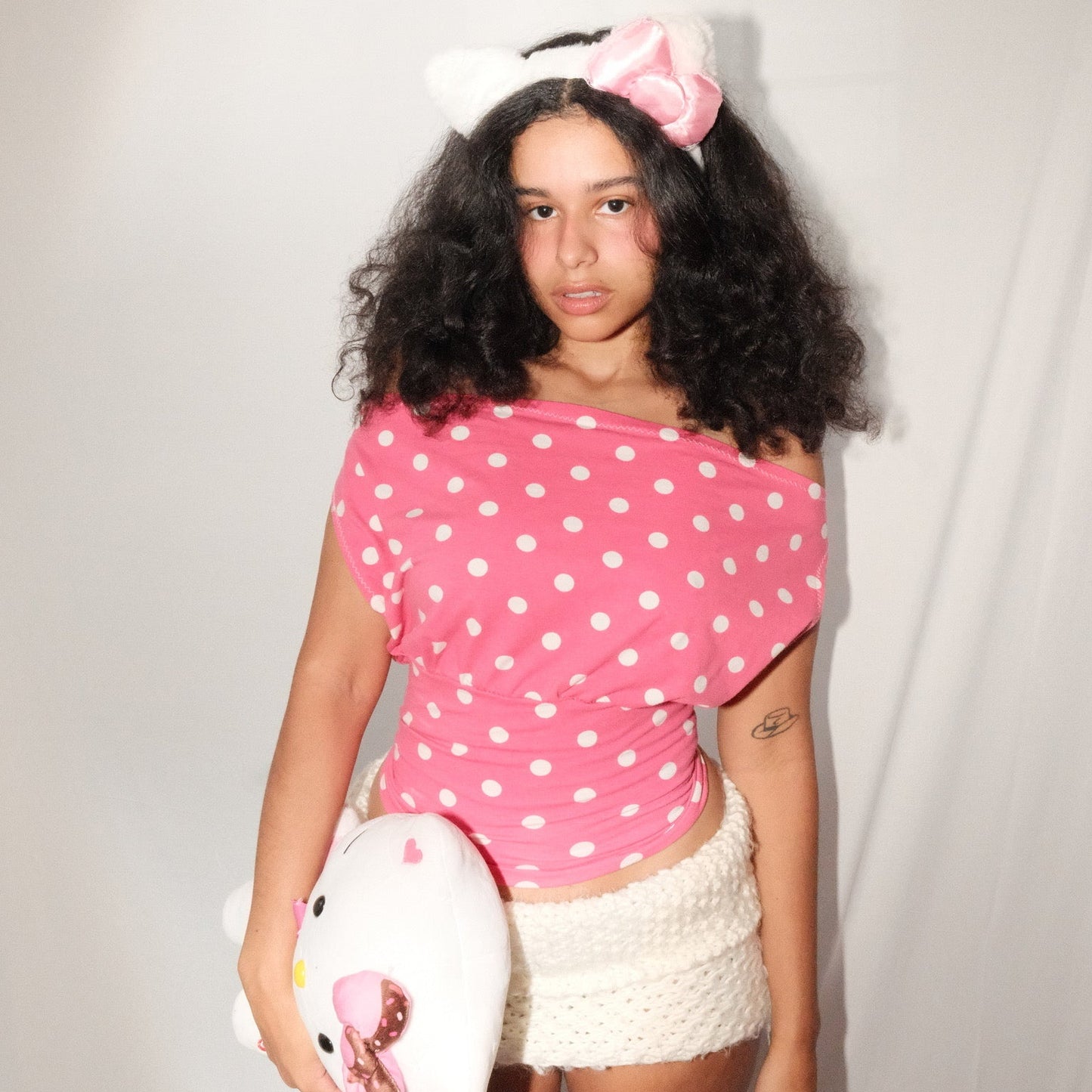 Person wearing a pink polka dot top and white shorts, holding a plush toy with a white background
