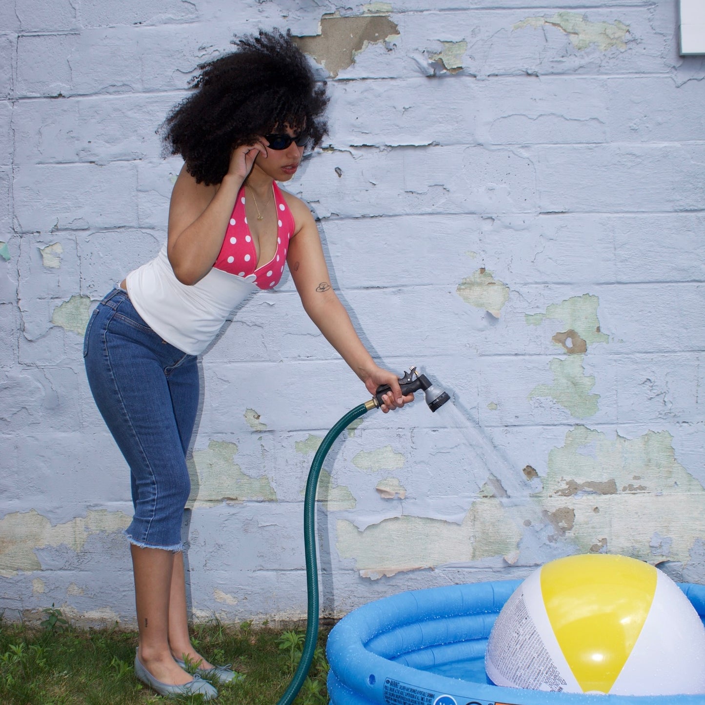 Woman using a hose to fill an inflatable pool with a beach ball inside, against a textured wall.