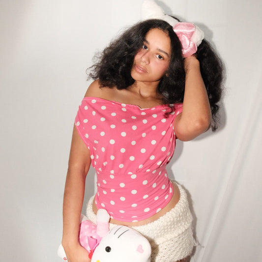 Woman wearing a pink polka dot top and white shorts, holding a Hello Kitty plush toy against a plain background.