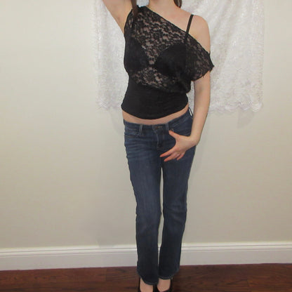 Person wearing a black lace top and blue jeans against a neutral wall.