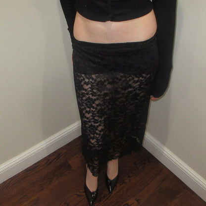 Person wearing a black lace skirt indoors against a plain wall.
