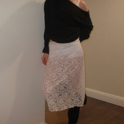 Person wearing a black top and white lace skirt against a plain wall.