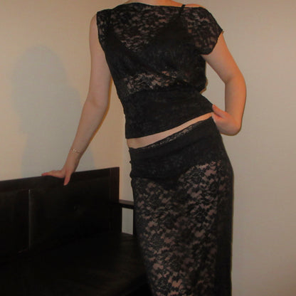 Person wearing a black lace outfit standing against a plain wall.