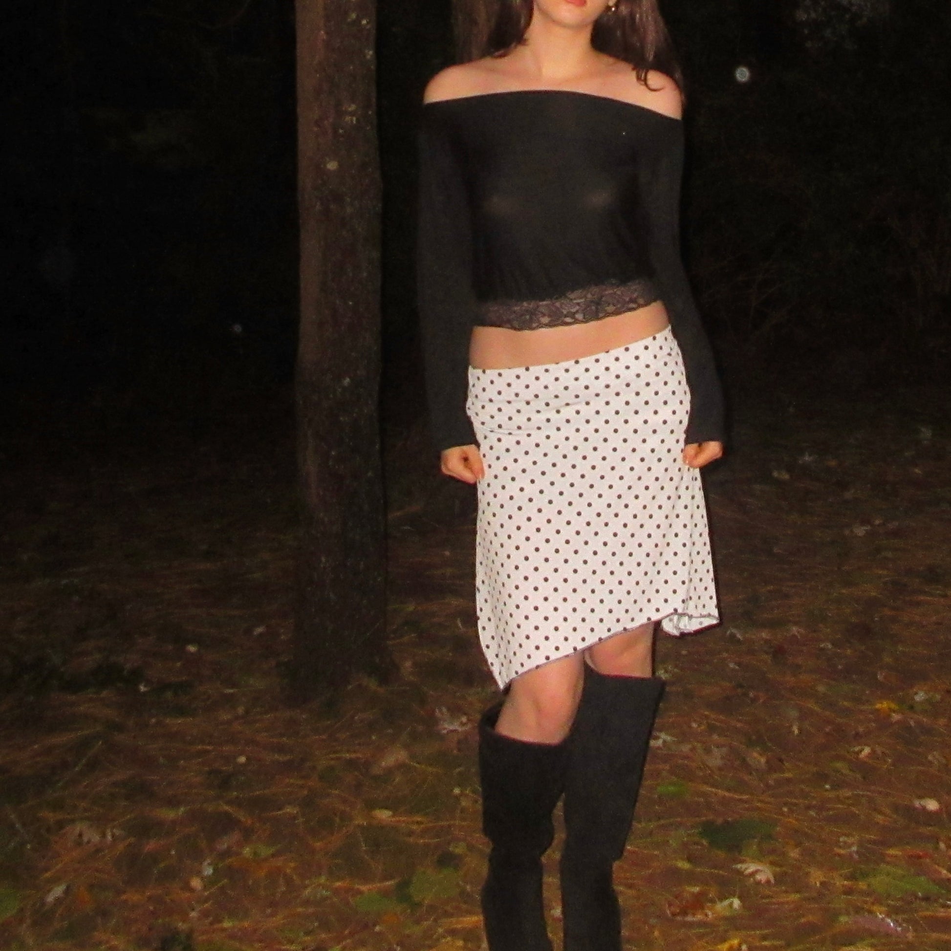 Person wearing a black top and polka dot skirt with knee-high boots in a dark forest setting.