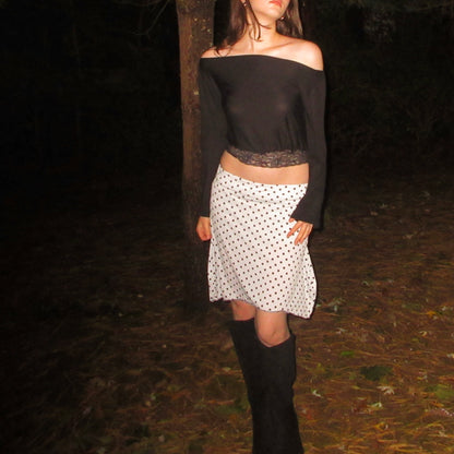 Person wearing a black off-shoulder top and white polka dot skirt in a dark forest setting.